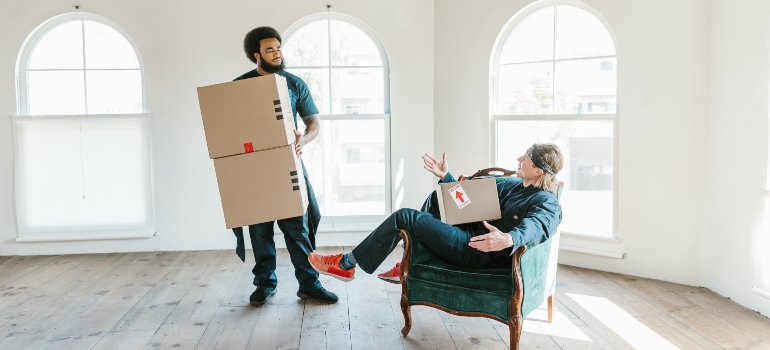 Two men, who are working for San Diego to Portland movers, chatting in the empty room while holding carton boxes.